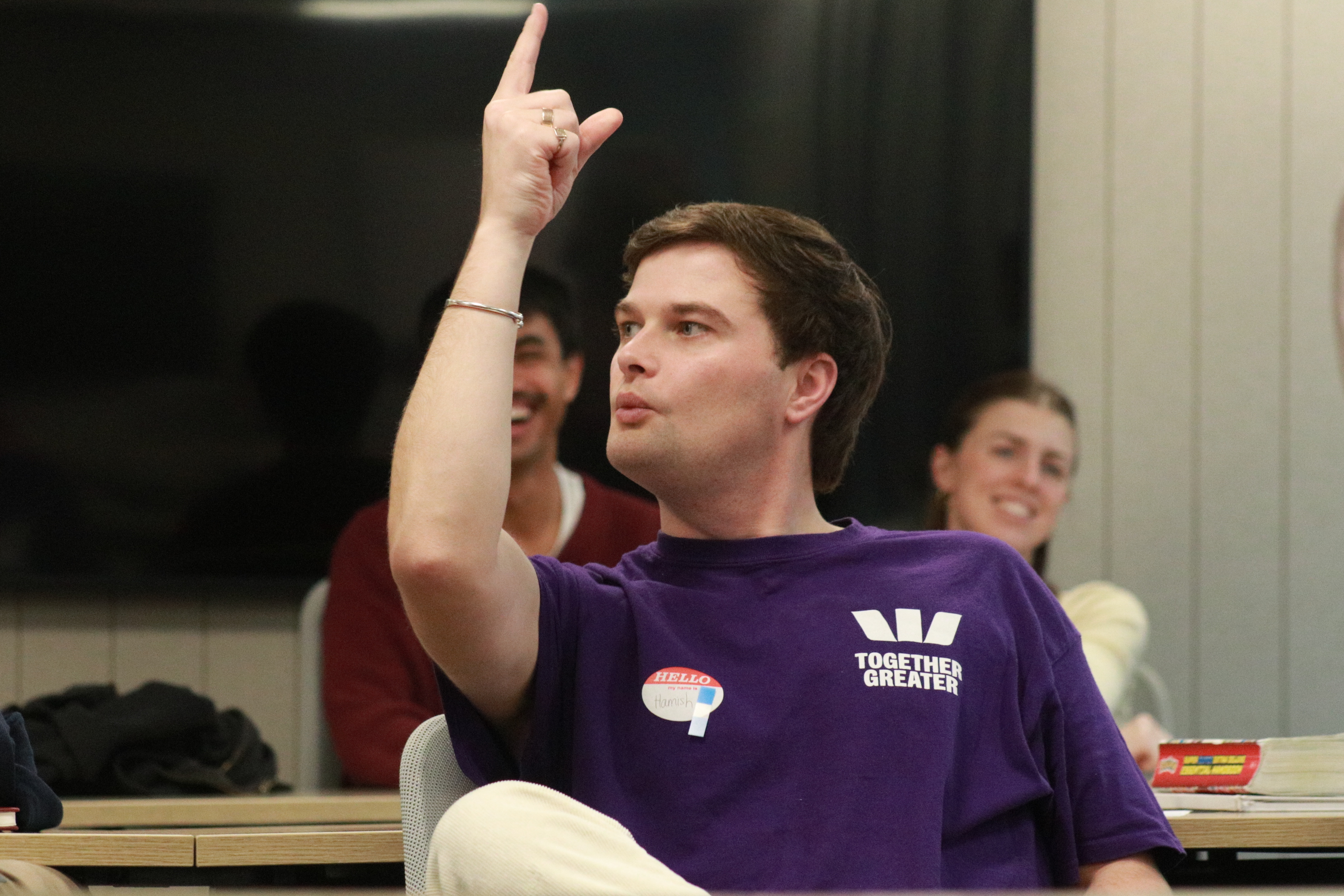 A person in a purple 'Together Greater' T-shirt sits in a classroom or meeting room, raising one finger while speaking. They wear a 'Hello' name tag, while two smiling people sit blurred in the background.